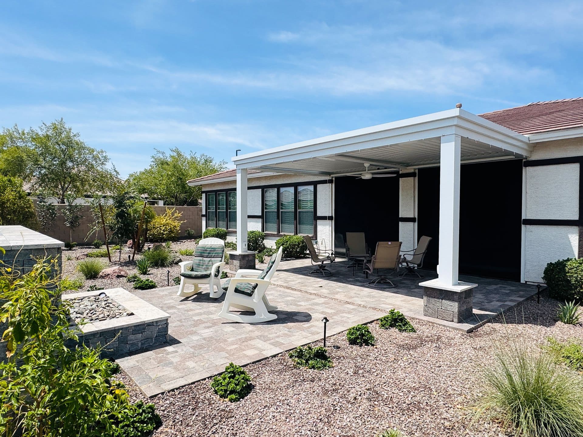 Modern backyard patio with white pergola, lounge chairs, dining seating, paved terrace, landscaped garden beds, and residential home under clear blue sky