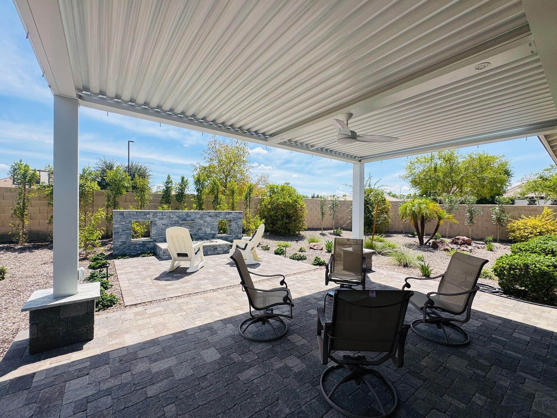Covered patio with metal roof and ceiling fan, featuring multiple lounge chairs around a fire pit, stone bench seating, and landscaped backyard with plants and block walls